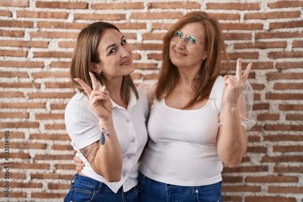 Hispanic mother and daughter wearing casual white t shirt smiling looking to the camera showing fingers doing victory sign. number two.