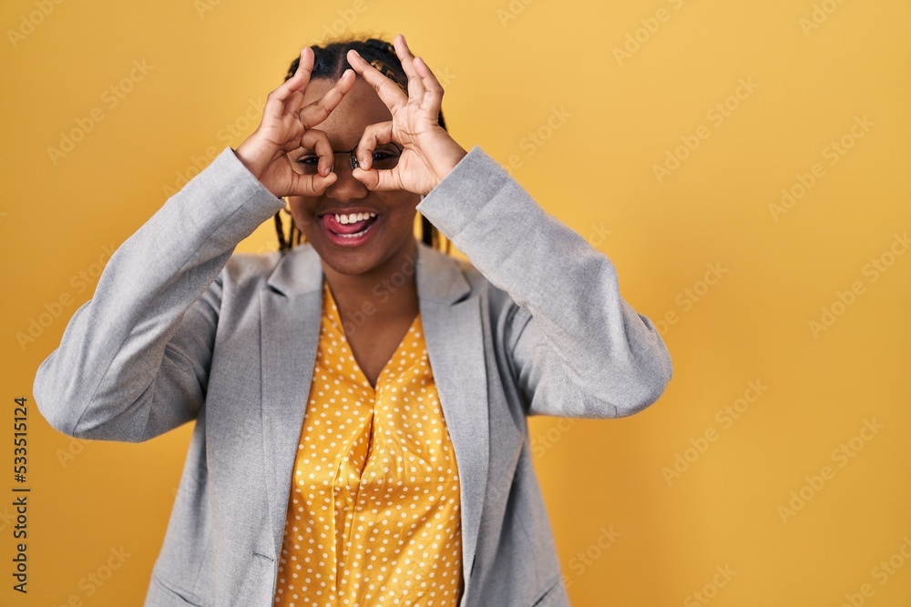 Fototapeta premium African american woman with braids standing over yellow background doing ok gesture like binoculars sticking tongue out, eyes looking through fingers. crazy expression.