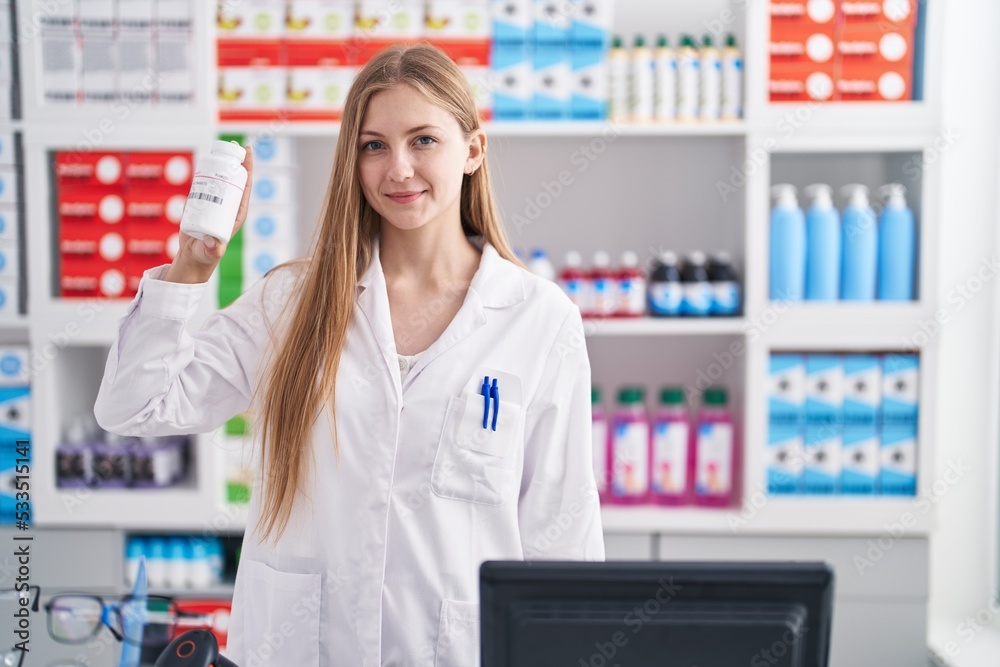 Young caucasian woman pharmacist smiling confident holding pills bottle at pharmacy