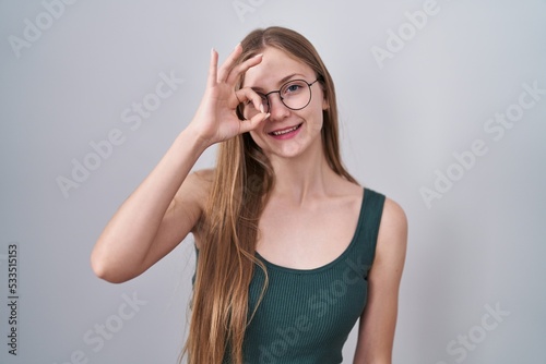 Young caucasian woman standing over white background doing ok gesture with hand smiling, eye looking through fingers with happy face.