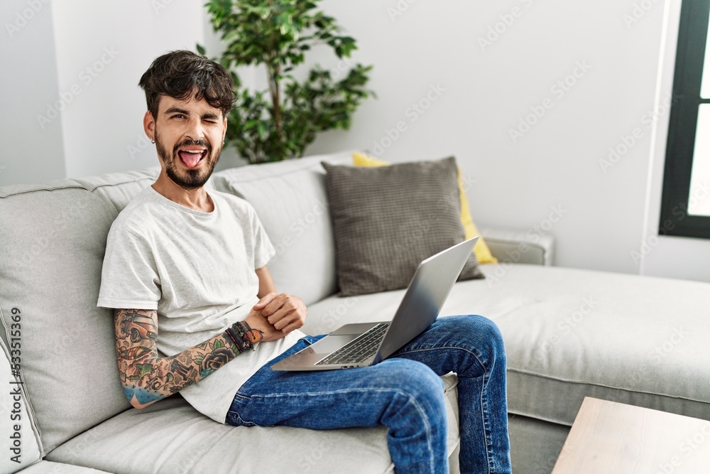 Hispanic man with beard sitting on the sofa sticking tongue out happy with funny expression. emotion concept.