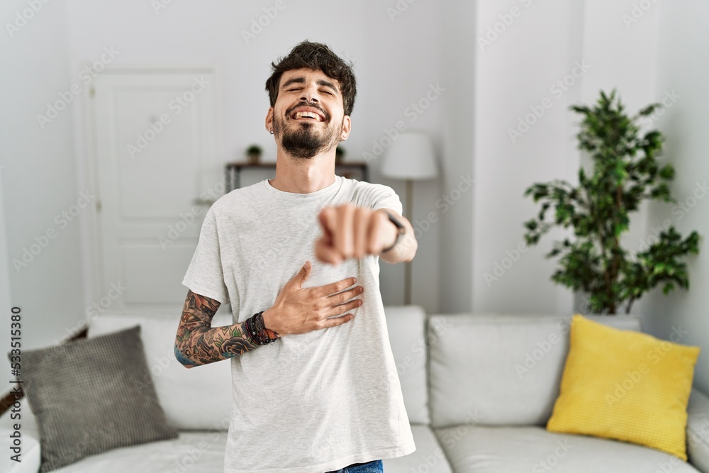Hispanic man with beard at the living room at home laughing at you, pointing finger to the camera with hand over body, shame expression