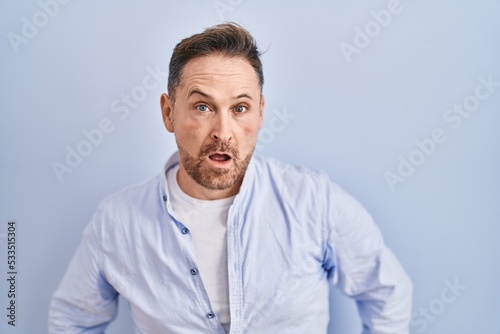 Middle age caucasian man standing over blue background in shock face, looking skeptical and sarcastic, surprised with open mouth © Krakenimages.com