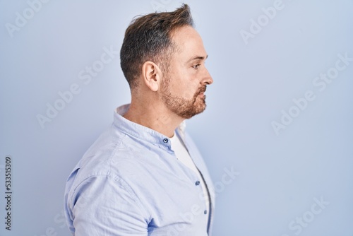 Middle age caucasian man standing over blue background looking to side, relax profile pose with natural face and confident smile.