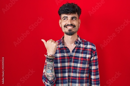 Young hispanic man with beard standing over red background smiling with happy face looking and pointing to the side with thumb up.