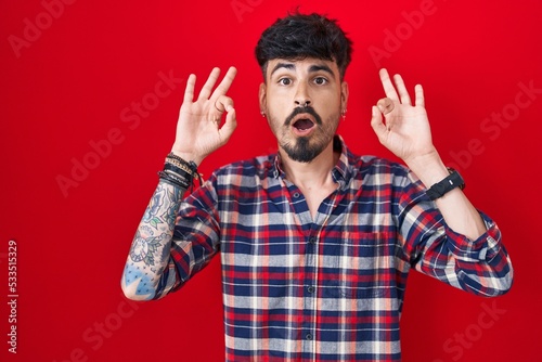 Young hispanic man with beard standing over red background looking surprised and shocked doing ok approval symbol with fingers. crazy expression