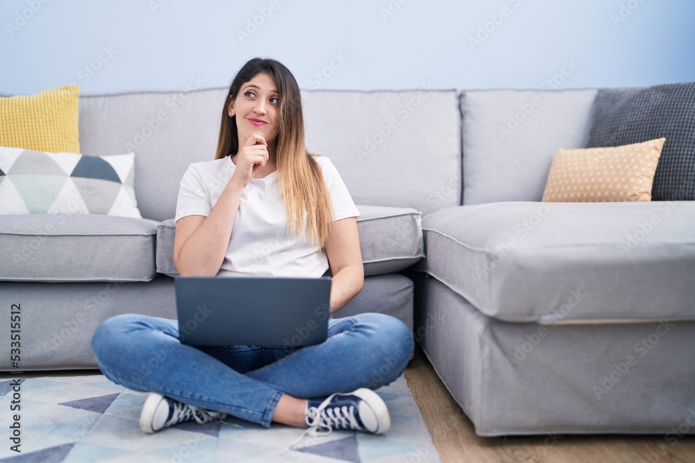 Obraz premium Young brunette woman sitting on the floor at home using laptop serious face thinking about question with hand on chin, thoughtful about confusing idea