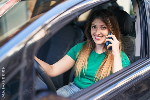 Young hispanic woman talking on the smartphone sitting on car at street