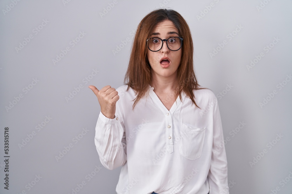 Brunette woman standing over white isolated background surprised pointing with hand finger to the side, open mouth amazed expression. Brunette woman standing over white isolated background surprised pointing with hand finger to the side, open mouth amazed expression.