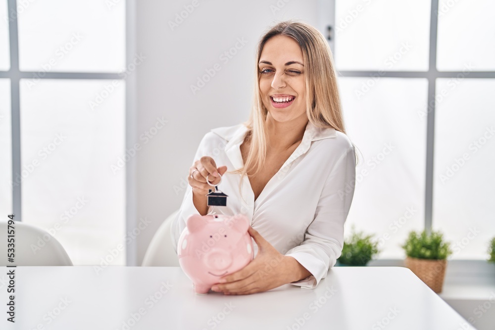 Young blonde woman holding piggy bank and house keys winking looking at the camera with sexy expression, cheerful and happy face. Young blonde woman holding piggy bank and house keys winking looking at the camera with sexy expression, cheerful and happy face.