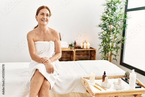 Middle age caucasian woman wearing towel sitting on massage board at beauty center