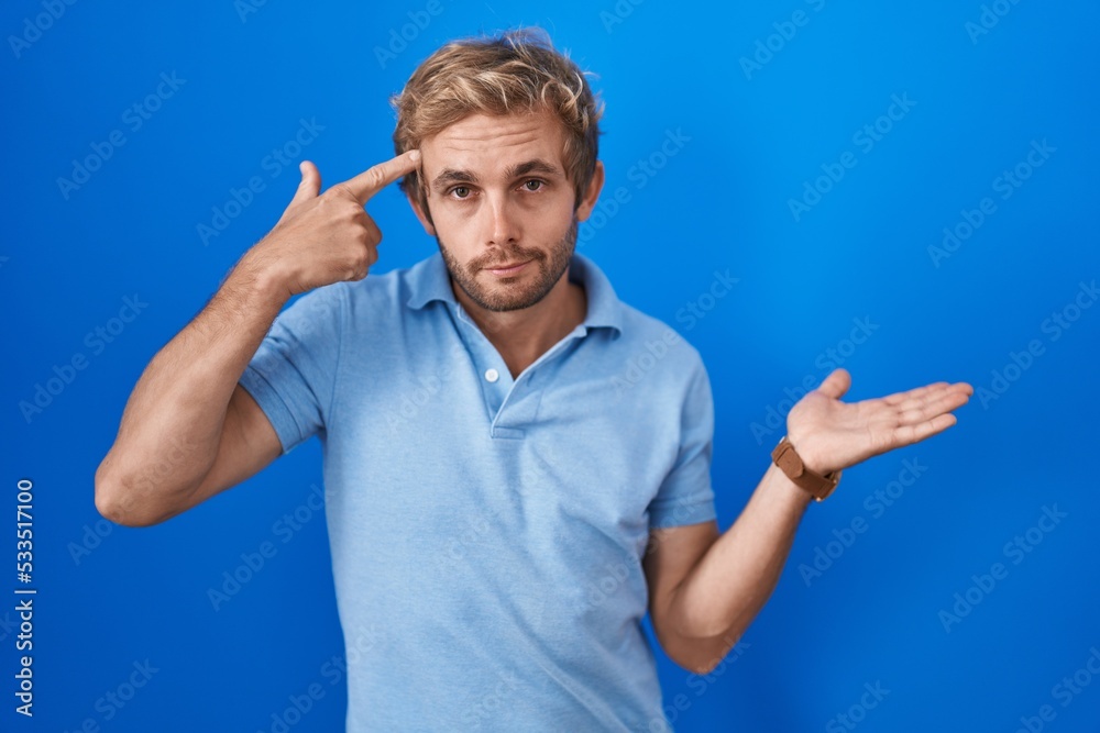 Caucasian man standing over blue background confused and annoyed with open palm showing copy space and pointing finger to forehead. think about it.