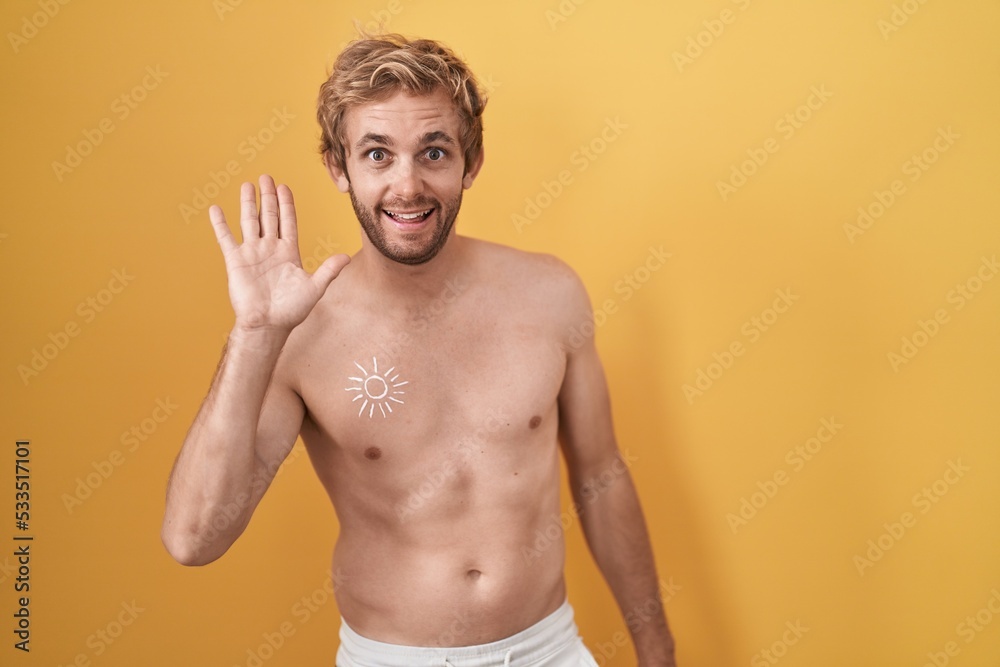 Caucasian man standing shirtless wearing sun screen waiving saying hello happy and smiling, friendly welcome gesture