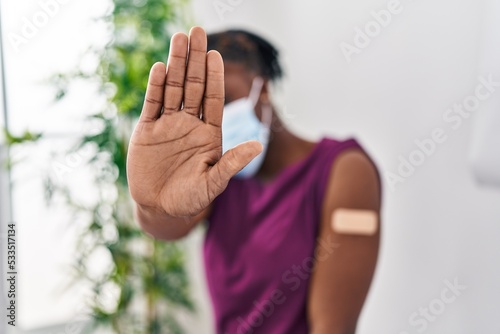 Beautiful black woman getting vaccine showing arm with band aid with open hand doing stop sign with serious and confident expression, defense gesture