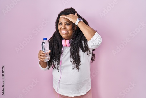 Plus size hispanic woman wearing sportswear and headphones very happy and smiling looking far away with hand over head. searching concept.