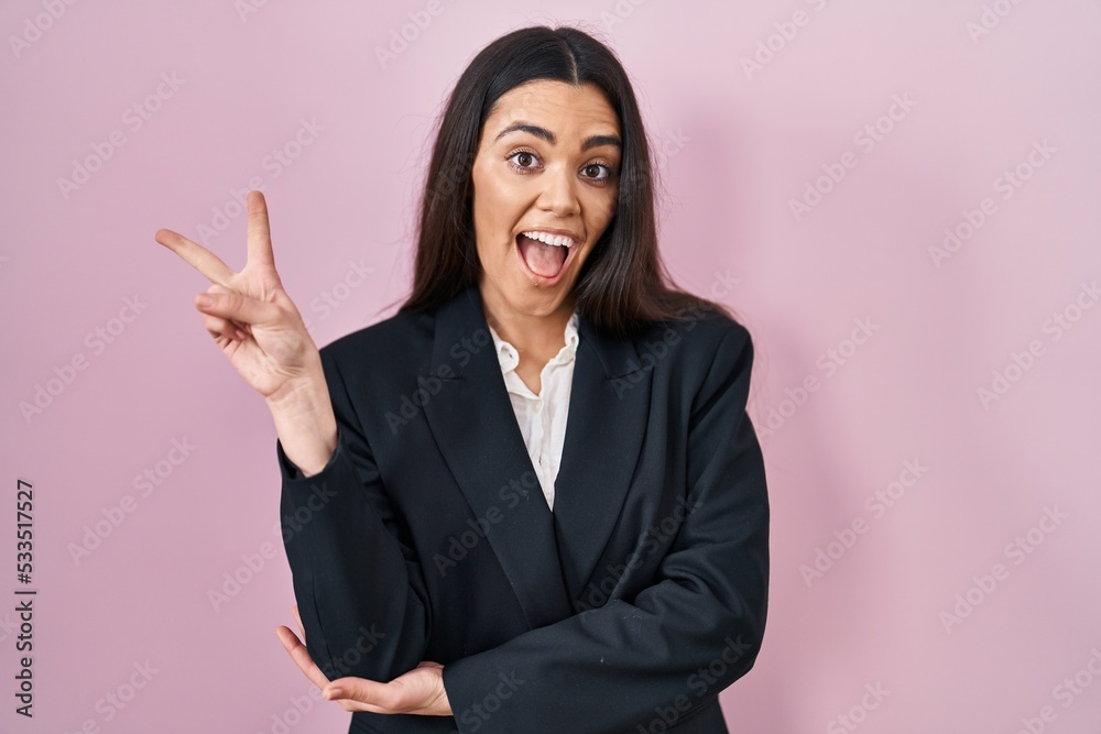 Young brunette woman wearing business style over pink background smiling with happy face winking at the camera doing victory sign with fingers. number two. Young brunette woman wearing business style over pink background smiling with happy face winking at the camera doing victory sign with fingers. number two.