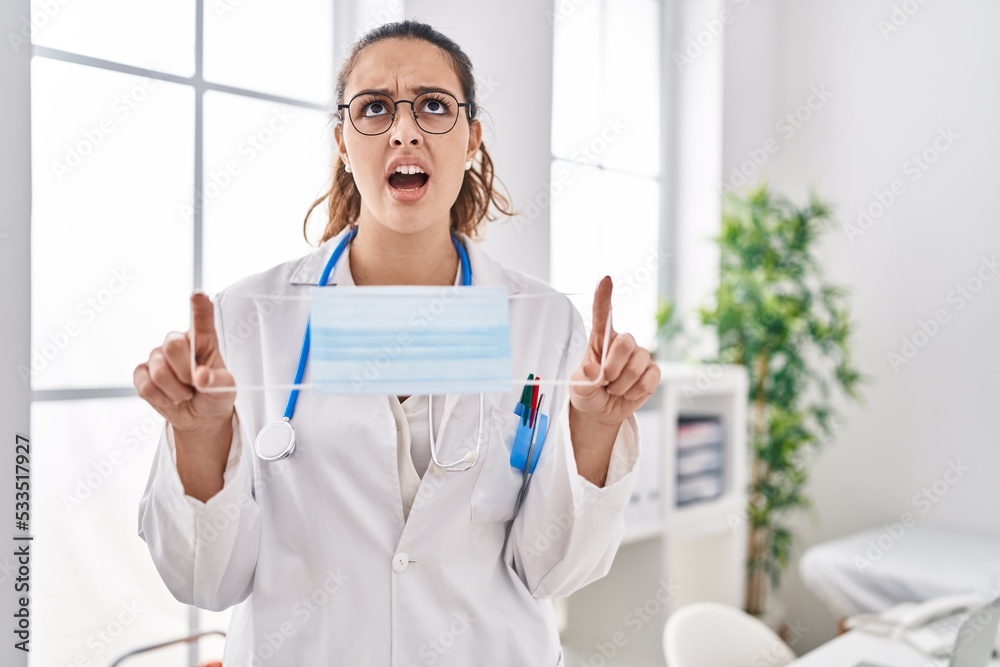 Young hispanic doctor woman holding safety mask angry and mad screaming frustrated and furious, shouting with anger looking up. Young hispanic doctor woman holding safety mask angry and mad screaming frustrated and furious, shouting with anger looking up.