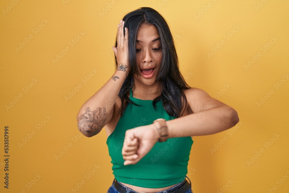 Brunette woman standing over yellow background looking at the watch time worried, afraid of getting late Brunette woman standing over yellow background looking at the watch time worried, afraid of getting late