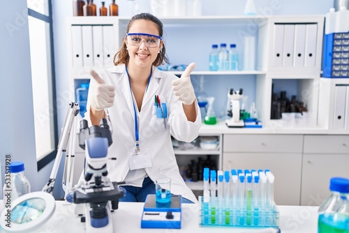 Young hispanic woman working at scientist laboratory approving doing positive gesture with hand  thumbs up smiling and happy for success. winner gesture.