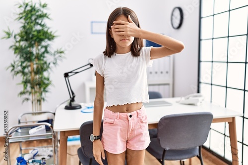 Young hispanic girl standing at pediatrician clinic covering eyes with hand, looking serious and sad. sightless, hiding and rejection concept