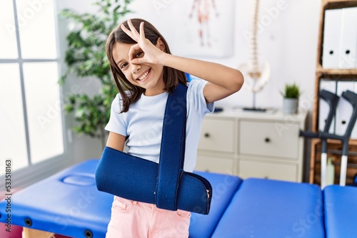 Young hispanic girl wearing arm on sling at rehabilitation clinic smiling happy doing ok sign with hand on eye looking through fingers photo