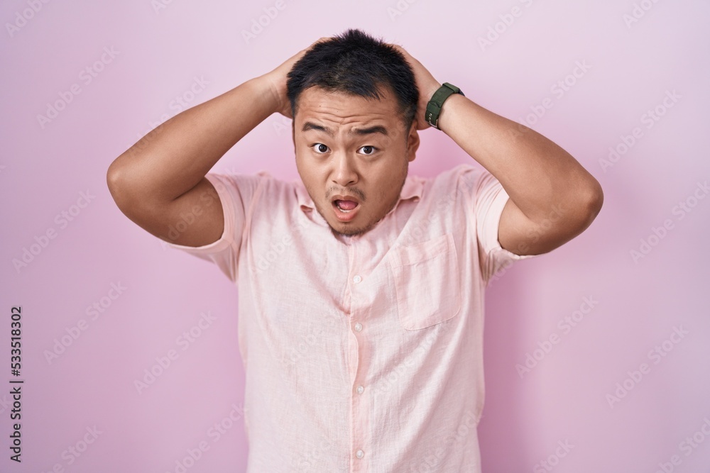 Chinese young man standing over pink background crazy and scared with hands on head, afraid and surprised of shock with open mouth