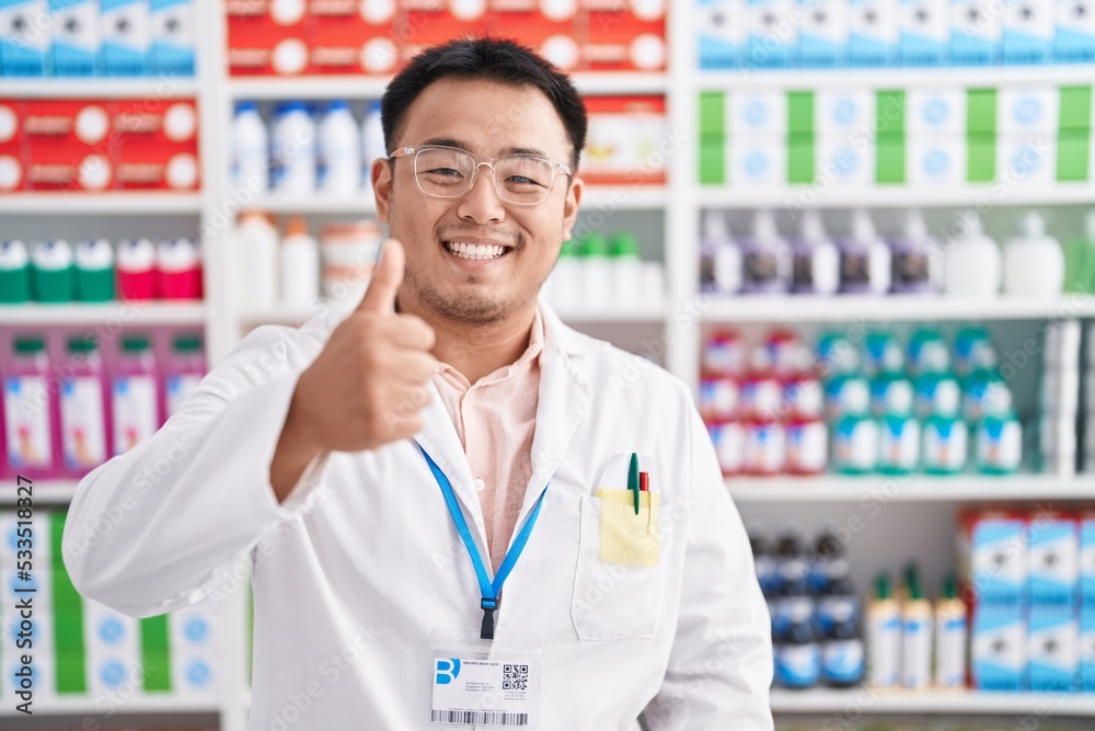 Chinese young man working at pharmacy drugstore doing happy thumbs up gesture with hand. approving expression looking at the camera showing success. Chinese young man working at pharmacy drugstore doing happy thumbs up gesture with hand. approving expression looking at the camera showing success.