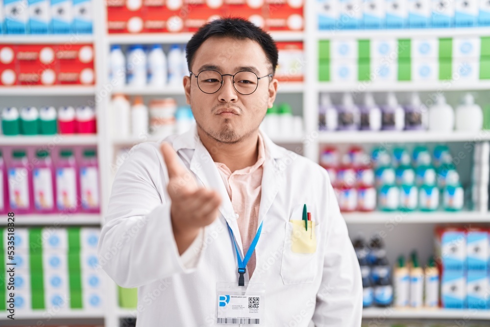 Chinese young man working at pharmacy drugstore looking at the camera blowing a kiss with hand on air being lovely and sexy. love expression.