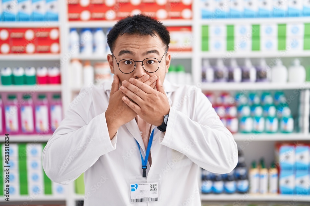 Chinese young man working at pharmacy drugstore shocked covering mouth with hands for mistake. secret concept.