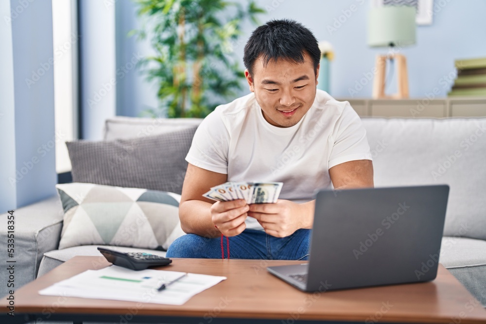 Young chinese man counting dollars sitting on sofa at home Young chinese man counting dollars sitting on sofa at home