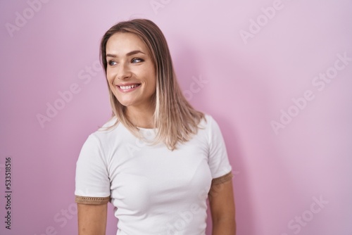 Blonde caucasian woman standing over pink background looking away to side with smile on face, natural expression. laughing confident.