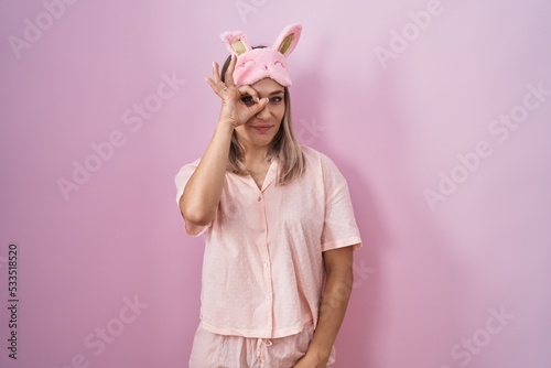 Blonde caucasian woman wearing sleep mask and pajama doing ok gesture with hand smiling, eye looking through fingers with happy face.