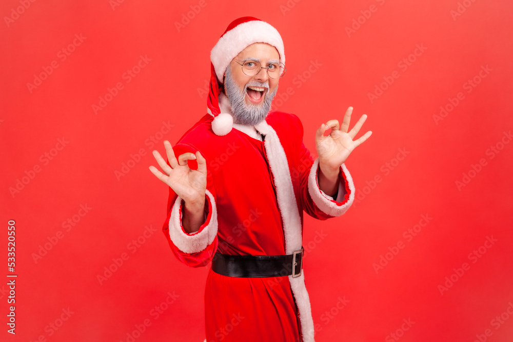 Portrait of happy elderly man with gray beard wearing santa claus costume with smile making ok gesture being agreeable, showing okay sign. Indoor studio shot isolated on red background.