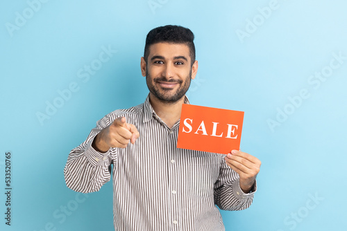 Photography Portrait of smiling satisfied businessman pointing at camera and holding card with sale inscription, low prices on Black Friday, wearing striped shirt