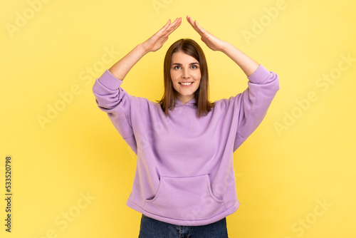 I'm in safety. Portrait of happy protected woman raising hands above head, making roof gesture, insurance, wearing purple hoodie. Indoor studio shot isolated on yellow background. © khosrork