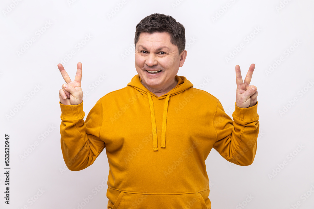 Fototapeta premium Happy positive middle aged man showing v sign to camera, winking and smiling, male wishing good luck, peace, wearing urban style hoodie. Indoor studio shot isolated on white background.