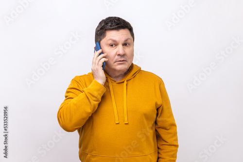 Serious man standing, talking smartphone, making call and having important conversation with friend or business partner, wearing urban style hoodie. Indoor studio shot isolated on white background.