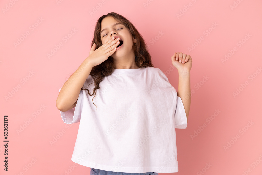 Portrait of adorable sleepy little girl wearing white T-shirt standing and yawning with closed eyes and raised arm, covering mouth with palm. Indoor studio shot isolated on pink background. Portrait of adorable sleepy little girl wearing white T-shirt standing and yawning with closed eyes and raised arm, covering mouth with palm. Indoor studio shot isolated on pink background.