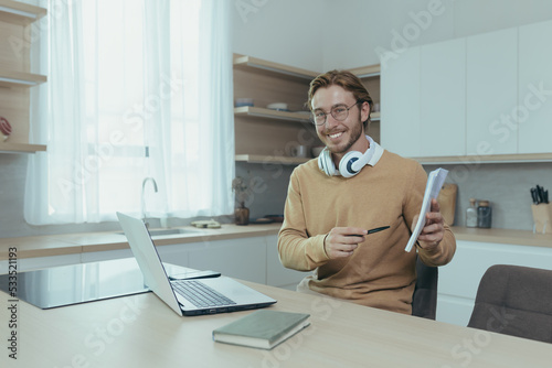 Young teacher teaches remotely from home, man works in kitchen at home, uses laptop for online call with communicates with students smiling explains assignment shows notebook.