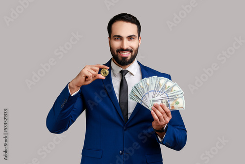 Man holding in hands and showing dollar bills and golden coin of crypto currency, transferring money into digital bitcoins, wearing official style suit. Indoor studio shot isolated on gray background.
