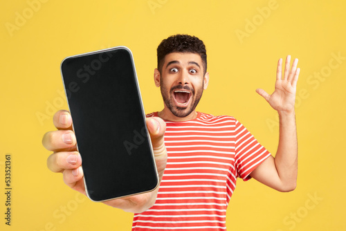 Amazed cheerful man with beard wearing red T-shirt showing mobile phone with blank display, expressing happiness, raised arm and screaming happily. Indoor studio shot isolated on yellow background.