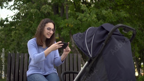 A young mother takes a picture on a smartphone of a child lying in a stroller. Woman in glasses with a phone in the park.