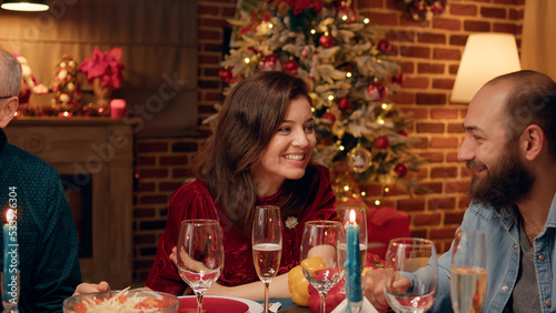 Festive wife talking with husband while celebrating Christmas at evening dinner family table. Pretty woman smiling heartily at camera while enjoying winter holidays with close family members.