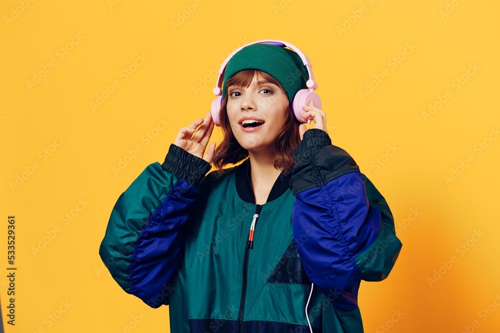a happy, joyful woman in a green jacket and hat enjoys music standing in pink headphones on a yellow background holding them with her hands and looking at the camera a happy, joyful woman in a green jacket and hat enjoys music standing in pink headphones on a yellow background holding them with her hands and looking at the camera