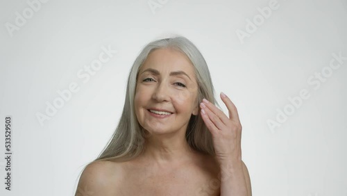 Beautiful senior grey haired lady doing anti wrinkles facial massage, smiling to camera, white studio background photo