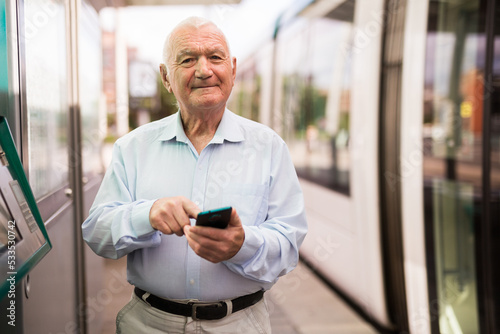Old man standing on tram station and using smartphone while waiting.