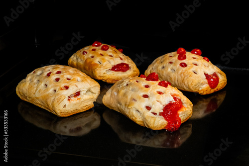 Four baked puff pastry buns with red strawberry jam on tray in electric oven, black background. Homemade bakery, food, cooking and pastry concept