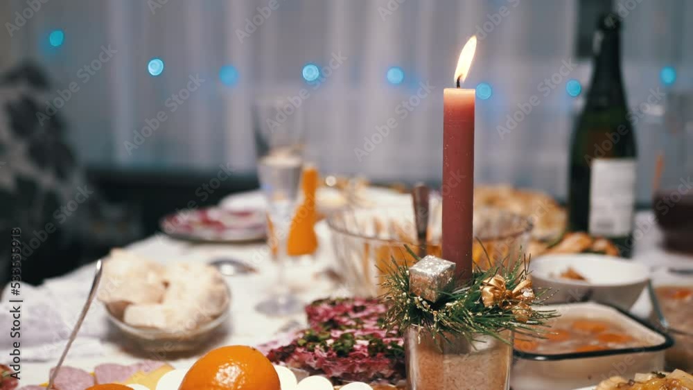 Burning Red Christmas Candle Burns with a Bright Flame on the New Year's Table. Christmas decorated table on eve of holiday on background bulbs garlands. Cozy family dinner in a festive atmosphere. Burning Red Christmas Candle Burns with a Bright Flame on the New Year's Table. Christmas decorated table on eve of holiday on background bulbs garlands. Cozy family dinner in a festive atmosphere.