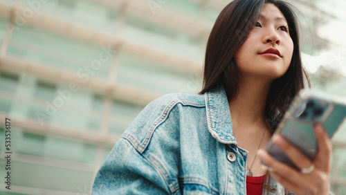 Portrait of long haired Asian girl texting on smartphone outdoors. Beautiful woman dressed casual using mobile phone on street