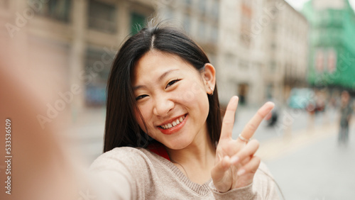 Fotografie, Obraz Cute Asian girl taking self portrait showing peace sign during walk through city